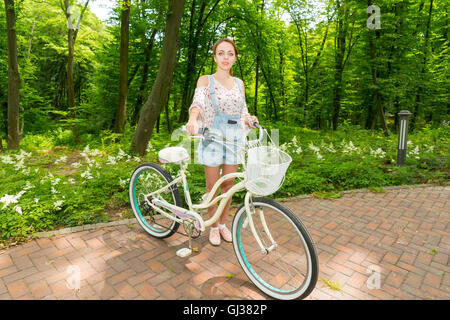 Glückliche Mädchen tragen Denim-Overalls und steh-locker geschnittene Bluse mit ihrem Fahrrad auf Ziegel in einem park Stockfoto