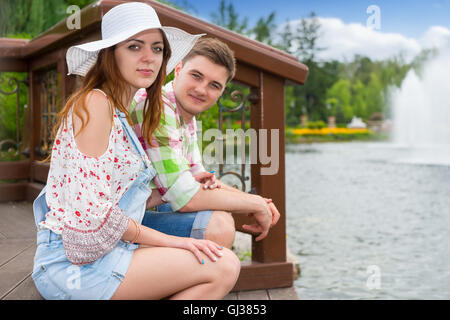 Paar sitzt auf dem Holzdeck gegenüber dem künstlichen See in einem Park mit Brunnen und verschiedene Bäume im Hintergrund Stockfoto