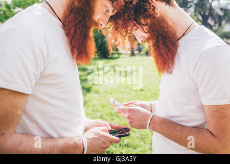 Junge männliche Hipster Zwillinge mit roten Bärten gegenüber einander SMS auf smartphones Stockfoto
