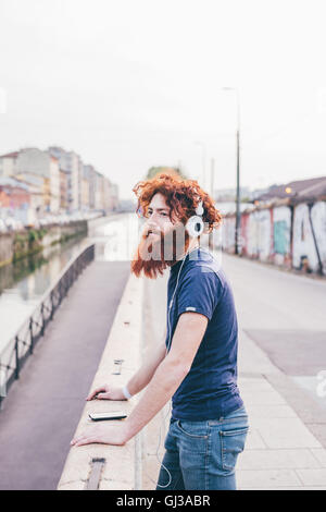 Junge männliche Hipster mit roten Haaren und Bart hören Kopfhörer auf Stadtbrücke Stockfoto
