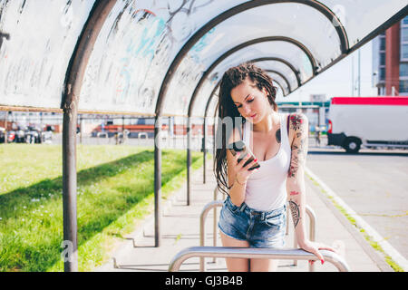 Junge Frau mit Dreadlocks lesen Smartphone in städtische Wartehäuschen Stockfoto