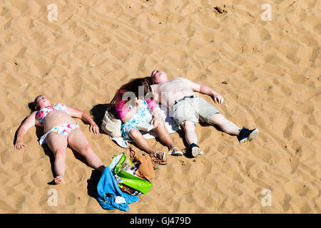 England, Broadstairs. Strand. Zwei Menschen, Mann und Frau, gekleidet Überdrehen Sonnenanbeter Verlegung in der Sonne auf dem Rücken mit einem dritten dazwischen sitzen, ein Buch zu lesen. Stockfoto