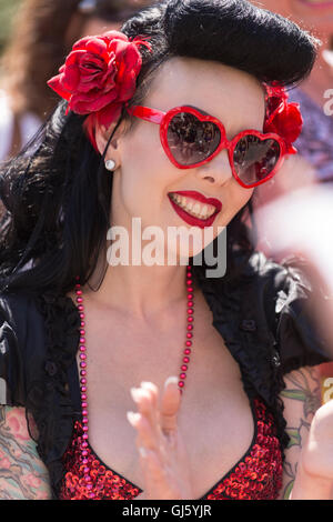 Frau mit schwarzen Haaren, roten Lippen, rote Blumen in ihrem Haar und roten Herzen geformt Sonnenbrille, lächelnd. Stockfoto