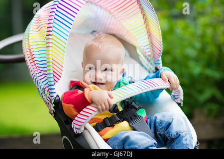 Baby Junge im weißen Pullover sitzen in weißen Kinderwagen auf einen Spaziergang in einem Park. Kind im regenbogenfarbenen Buggy. Stockfoto