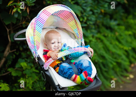 Baby Junge im weißen Pullover sitzen in weißen Kinderwagen auf einen Spaziergang in einem Park. Kind im regenbogenfarbenen Buggy. Stockfoto