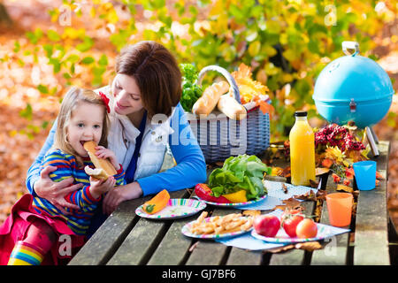 Glückliche junge Mutter mit kleinen Mädchen Grillen von Fleisch und Sandwich und Salat auf einem Picknicktisch im sonnigen Herbst Park. Stockfoto