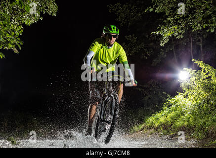 Mountainbiker bei Nacht Fahrt durch Wald Stream und spritzt Wasser herum. Frontansicht Stockfoto