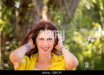 elegant und glamourös Reife Frau mit bunten Armbänder und gelben Schal ist in einem grünen Park lächelnd, während des Spielens mit braunen Haaren Stockfoto