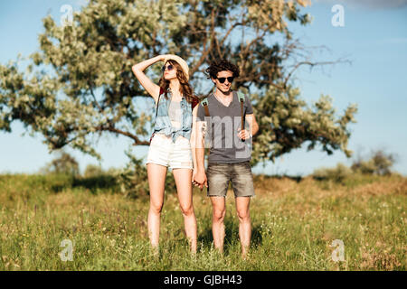 Junge attraktive Hipster paar stehend gegen Bäume auf dem Grund und Hand in Hand Stockfoto