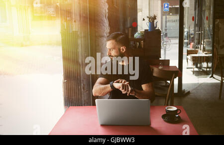 Junge bärtige Geschäftsmann tragen schwarze Tshirt arbeiten Laptop Urban Cafe.Man sitzen Holztisch Tasse Kaffee mit Blick durch Fenster Touch Smartwatch.Coworking Prozess Business Startup.Blurred Hintergrund Stockfoto