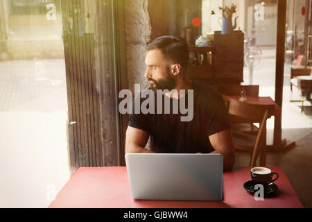 Young bärtigen Geschäftsmann tragen schwarz Tshirt arbeiten Laptop Urban Cafe.Man sitzen Holz Tisch Tasse Kaffee auf der Suche durch Window.Coworking Prozess Business Startup.Blurred Background.Sunlight Wirkung. Stockfoto
