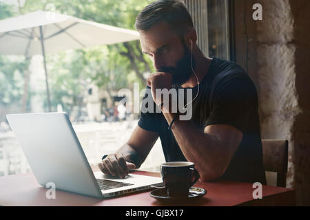 Konzentrierte junge bärtige Geschäftsmann tragen schwarze Tshirt arbeiten Laptop Urban Cafe.Man sitzen Holztisch Tasse Kaffee mit Blick über Window.Coworking Prozess Business Startup.Blurred Hintergrund Stockfoto