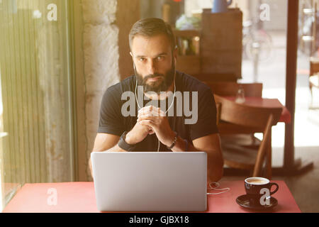 Junge Bearded Geschäftsmann tragen schwarz Tshirt arbeiten Laptop Urban Cafe.Man sitzen Holz Tisch Tasse Kaffee hören Music.Coworking Prozess Business Startup.Blurred Background.Sunlight Wirkung. Stockfoto