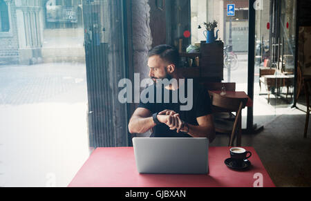 Junge bärtige Geschäftsmann tragen schwarze Tshirt Urban Cafe.Man Laptop sitzen Holztisch Tasse Kaffee mit Blick durch Fenster Touch Smartwatch.Coworking Business Startup.Color Prozessfilter arbeiten. Stockfoto