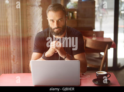 Junge bärtige Geschäftsmann tragen schwarze Tshirt arbeiten Laptop Urban Cafe.Man Holztisch Tasse Kaffee hören Music.Coworking Prozess Business Startup.Blurred Background.Sunlight Farbeffekt zu sitzen. Stockfoto