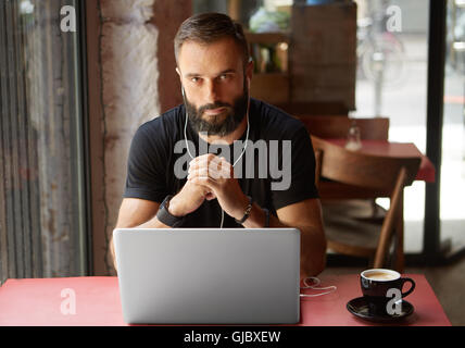 Hübscher Junge bärtige Geschäftsmann tragen schwarze Tshirt Urban Cafe.Man Laptop sitzen Holztisch Tasse Kaffee hören Music.Coworking Prozess Business Startup.Blurred Hintergrund arbeiten. Stockfoto