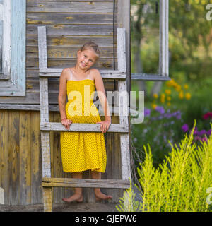 Niedliche kleine Mädchen steht auf einer Holzleiter an der Wand eines Dorfhauses. Stockfoto