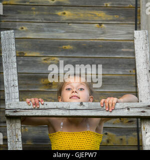 Niedliche kleine Mädchen steht auf einer Holzleiter an der Wand eines Dorfhauses. Stockfoto