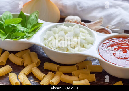 Basilikum, in Scheiben geschnittene Zwiebeln und Tomatenmark in weiße Schüssel mit rohen Nudeln und Knoblauch auf hölzernen Hintergrund verstreut. Konzept-ima Stockfoto