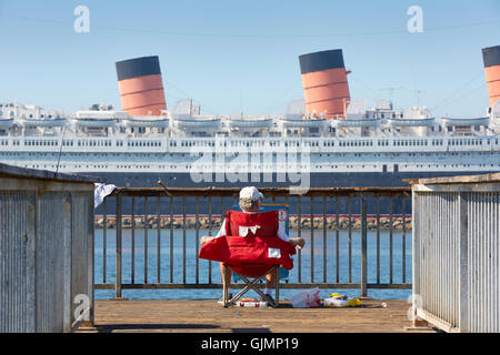 Mann, sitzend Strand Angeln von einem Pier mit Blick auf die RMS Queen Mary, lange Hafen. Stockfoto