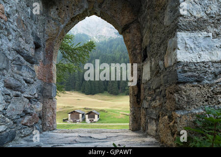 Blick durch das leere Tor Bogen einer alten Burgruine auf zwei Hütten. Grünen Wald und Wiese im Hintergrund sichtbar. Stockfoto