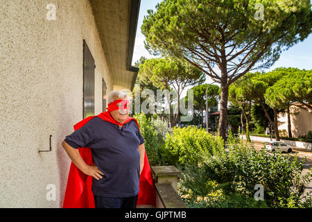 Lustige und lächelnd senior Mann posiert als Superheld mit roten Umhang und Maske hält stolz seine Hände auf den Hüften auf dem Balkon seines Hauses Stockfoto