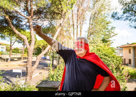 Lustige und fröhliche senior Mann posiert als Superheld mit roten Umhang und Maske hebt Arm vorbereiten, in einer ruhigen Wohngegend zu fliegen Stockfoto