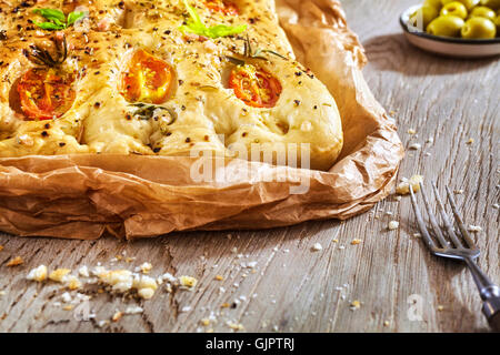 Frisch gebackene traditionelle italienische Focaccia mit Tomaten und Kräutern in Backpapier auf einem alten Holztisch. Stockfoto