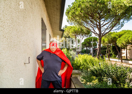 Lustige und lächelnd senior Mann posiert als Superheld mit roten Umhang und Maske hält stolz seine Hände auf den Hüften auf dem Balkon seines Hauses Stockfoto