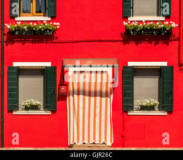 Dunkelgrüne Fensterläden an einer leuchtend rot gerenderten Außenwand des traditionellen Hauses auf der Insel Burano. Italien. Stockfoto