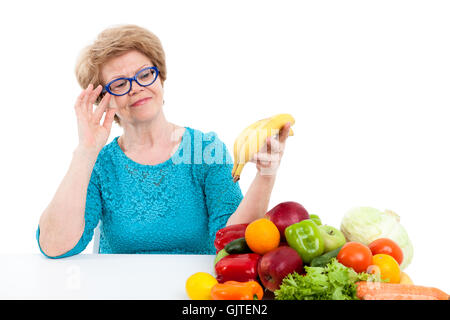 Ältere Frau mit Blick auf reife Banane in der hand, sitzen mit frischem Obst und Gemüse auf Tisch, isoliert auf weißem Hintergrund Stockfoto