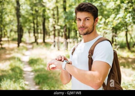 Lächelnd jungen Mann mit Rucksack und Uhr stehen im Wald Stockfoto