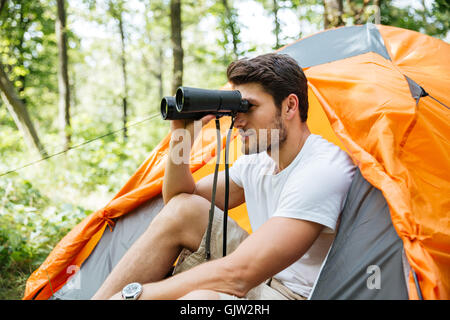 Attraktive junge Mann Tourist mit einem Fernglas sitzen in der Nähe von touristischen Zelt im Wald Stockfoto