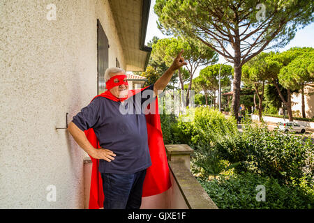 Lustige und fröhliche senior Mann posiert als Superheld mit roten Umhang und Maske hebt Arm vorbereiten, in einer ruhigen Wohngegend zu fliegen Stockfoto