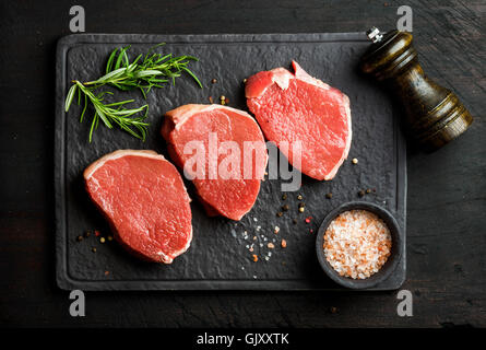 Rohes Rindfleisch Auge Runde Steaks mit Gewürzen und Rosmarin auf schwarzem Schiefer Steinplatte in dunklem Holz, Ansicht von oben, horizontal Stockfoto