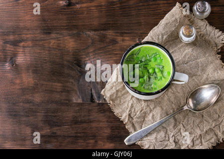 Weiße Vintage Tasse mit Creme von Erbsensuppe mit geschnittenem Basilikum serviert mit Löffel, Salz- und Pfefferstreuer auf Sackleinen über dunkle woode Stockfoto