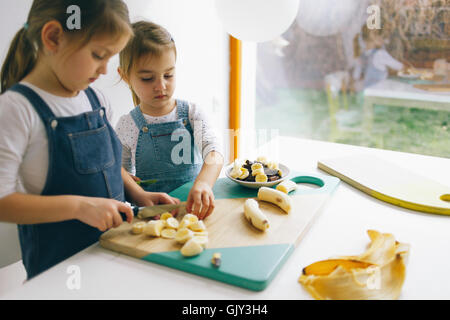 Zwei kleine Mädchen, die Banane auf dem Holzbrett auf dem Holzbrett zerkleinern Stockfoto