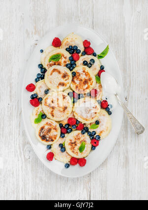 Syrniki oder Quark Pfannkuchen mit frischen Waldbeeren und saurer Sahne-Sauce im Teller über weißen hölzernen Hintergrund, Stockfoto