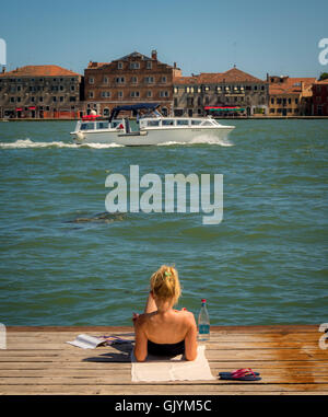 Junge Frau liegend auf einem Holzsteg über den Canale della Giudecca, mit einem Boot und in der Ferne die Insel Giudecca. V Stockfoto
