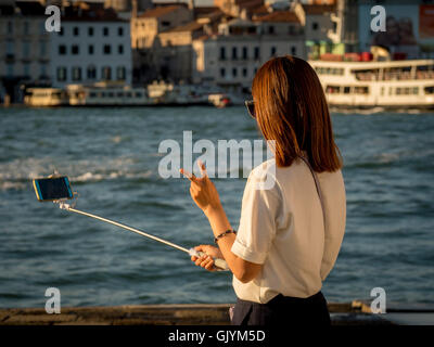 Asiatische Frau macht ein Selfie mit ihrem Smartphone und Selfie-Stick am Wasser in Venedig und macht mit ihrer Hand ein V- oder Friedenssymbol Stockfoto