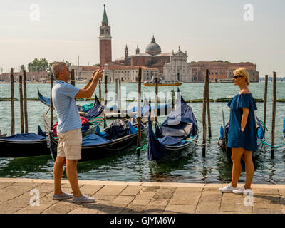 Männlichen Erwachsenen Touristen fotografieren Erwachsenfrau am Rand Wassers der Riva Degli Schiavoni, mit Ankern Gondeln und San Giorgio M Stockfoto