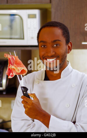 Glücklich Chef weiße Kleidung hält Fleischstücke mit großem Grill Gabel, glücklich lächelnd zu Kamera Stockfoto