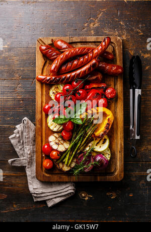 Gegrillte Würstchen und Gemüse auf Schneidebrett auf dunklem Holz Stockfoto