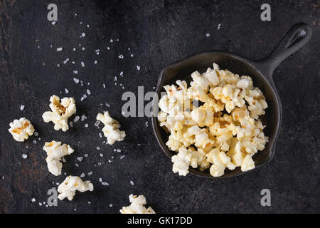 Vorbereitete gesalzene Popcorn serviert mit Meersalz in kleinen Eisen-Guss-Pfanne über schwarzes Eisen strukturiertem Hintergrund. Draufsicht mit Kopie spac Stockfoto