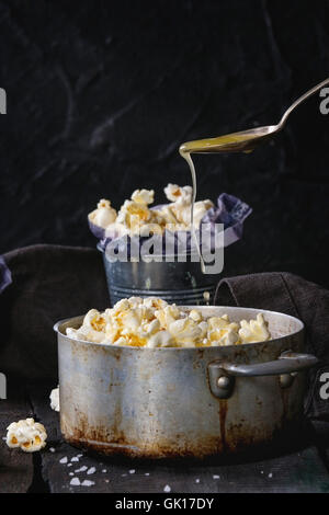 Vorbereitete gebutterte Popcorn mit Meersalz in kleinen Eimern und Vintage-Aluminium-Pfanne auf alten Holz Küchentisch serviert. Geschmolzen, aber Stockfoto