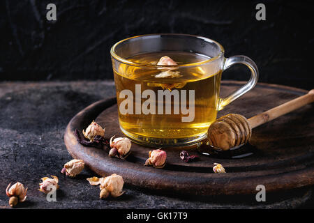 Glastasse heißer Kräutertee mit trockenen Rosen Knospen, serviert mit Honig auf Honig Dipper in dunklem Holz Schneidebrett über Blavk text Stockfoto