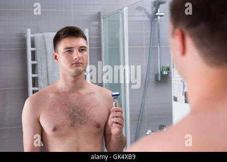 Mann nach der schlechten Rasur mit Pflaster auf seinem Gesicht ein Rasiermesser stehend vor dem Spiegel in das modern geflieste Bad zu Hause hält. Stockfoto