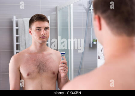 Hübscher junger Mann nach der schlechten Rasur mit Pflaster auf seinem Gesicht ein Rasiermesser stehend vor dem Spiegel in das modern geflieste Bad zu Hause hält. Stockfoto