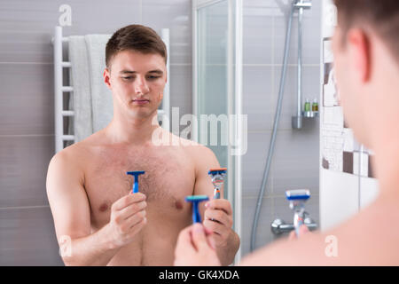 Hübscher junger Mann hält Rasierer und wählt das beste stehen im Vordergrund des Spiegels in das moderne geflieste Badezimmer zu Hause. Stockfoto