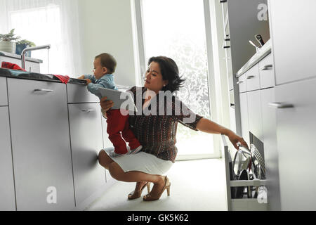 Mutter ist digital Tablet lesen, während ihr Baby auf ihren Händen, Küchenspüle erforscht Stockfoto